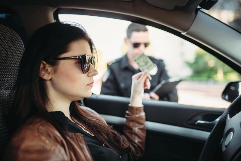 Vodička v slnečných okuliaroch sedí v aute a podáva vodičský preukaz policajtovi, ktorý jej píše pokutu za rýchlosť.